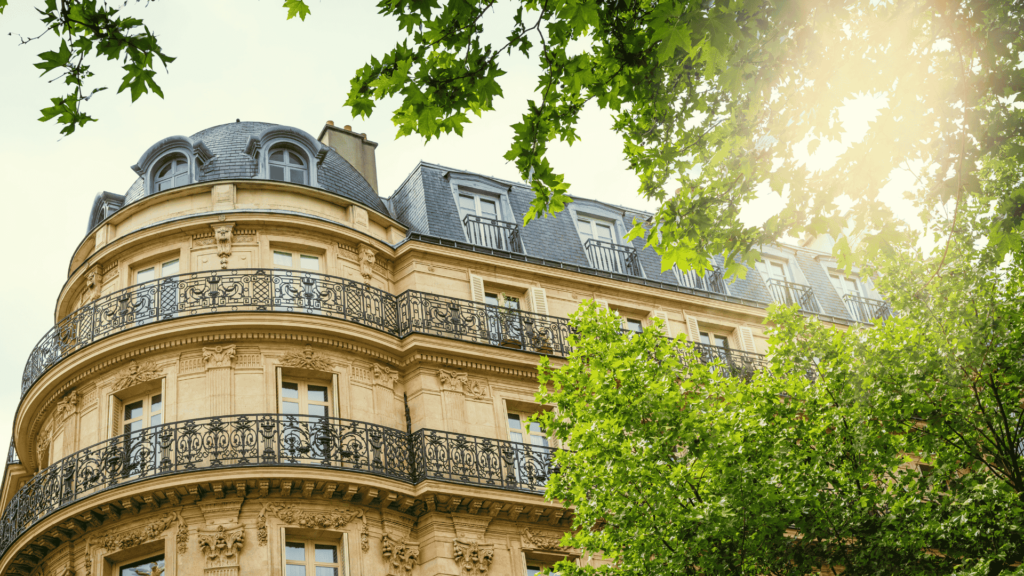 Elegant Parisian-style apartment building with ornate iron balconies, viewed through green trees in sunlight.