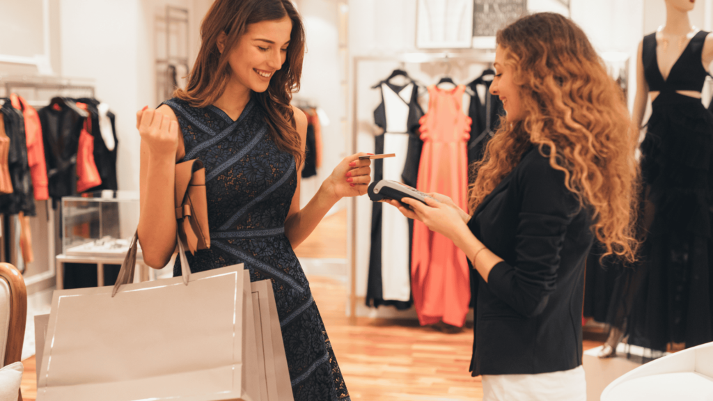 Smiling woman holding shopping bags pays with a card at a clothing store checkout.